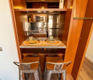 Een compacte keuken met een magnetron, fornuis en koelkast. Er zijn twee krukken en een kleine tafel met een decoratief bord.