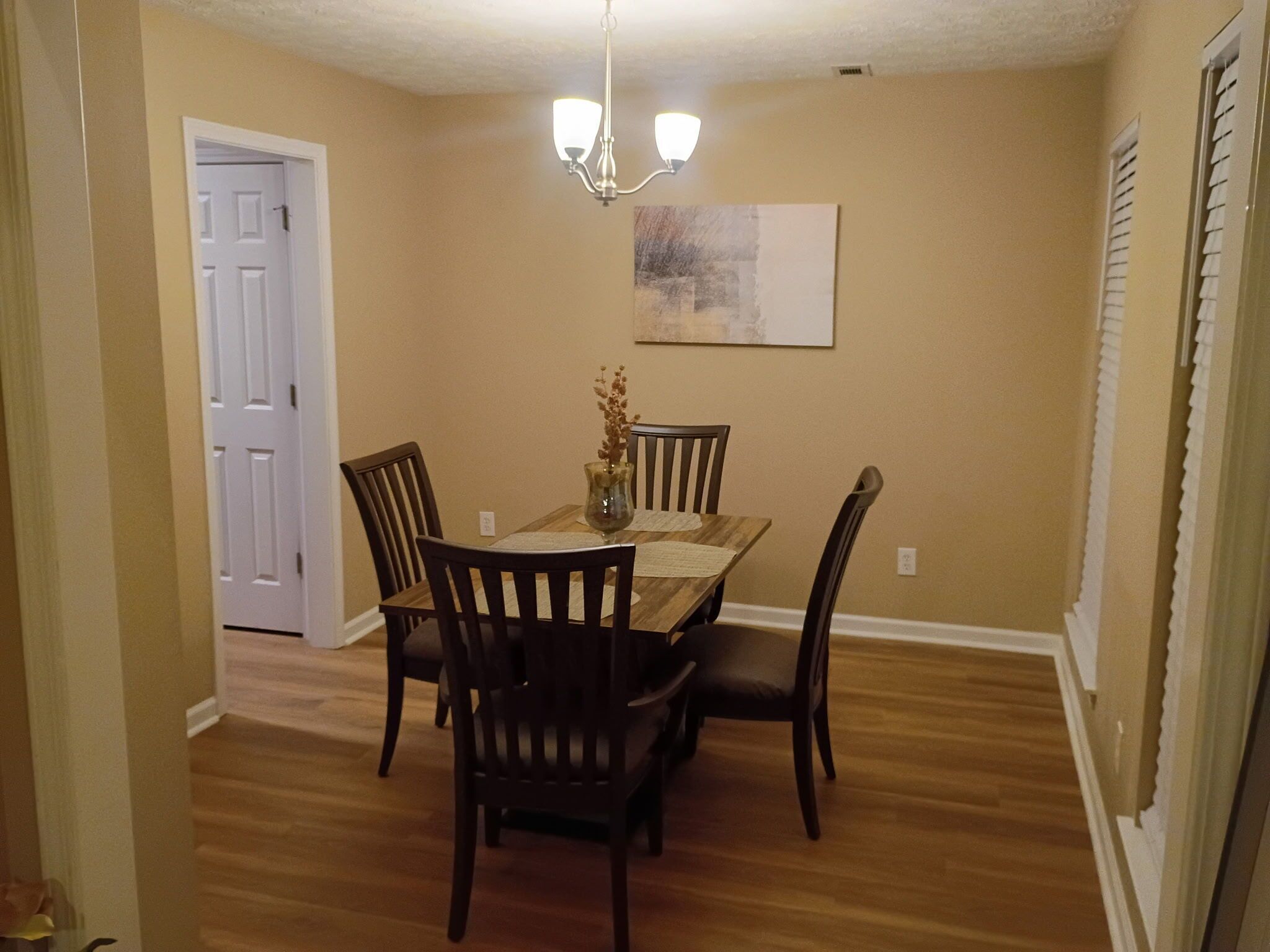 A dining room with a wooden table, four chairs, a chandelier, and a painting on the wall.