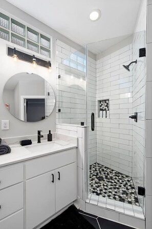 A modern bathroom with a glass-enclosed shower, white tiled walls, a round mirror, and a white vanity with a black faucet. - Gated, Modern & Spacious 3 BR House - 5 min drive to NRG Stadium - WiFi, Laundry (Houston)