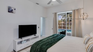 Une chambre avec un lit, une télévision, un ventilateur de plafond, une fenêtre avec des rideaux et une vue sur une piscine.