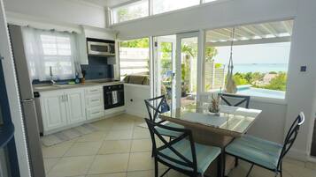 A kitchen with white cabinets, a microwave, and an oven. There is a dining table with chairs and a view of the ocean through large windows.