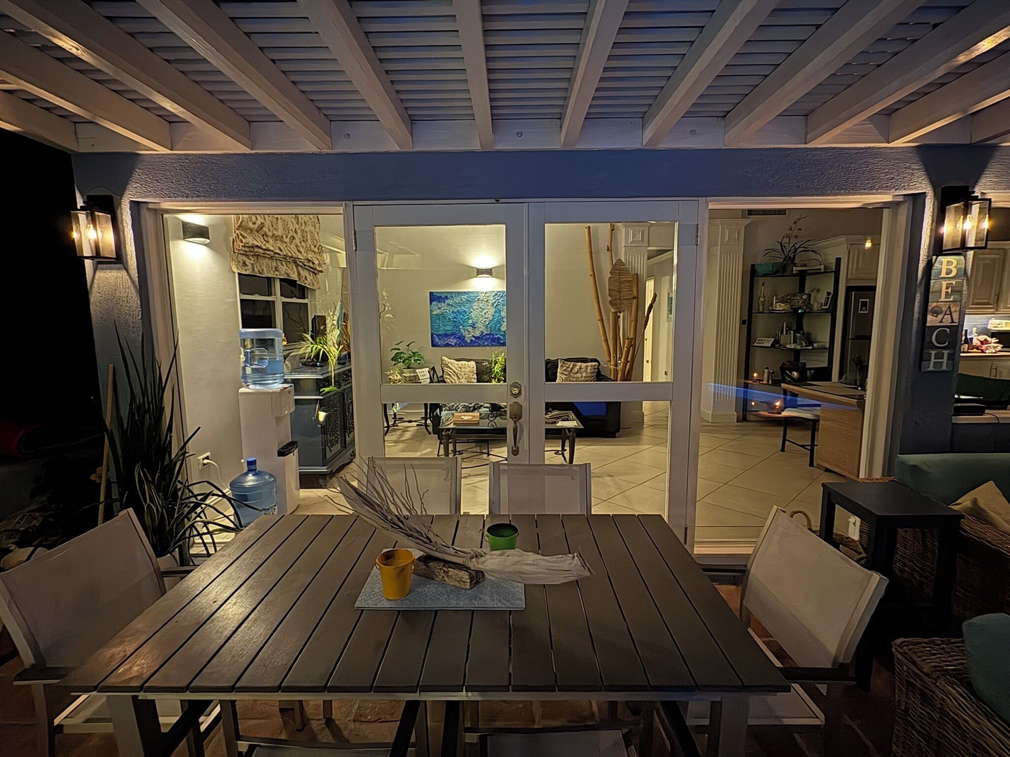 An outdoor dining area with a table and chairs, a water dispenser, and a view into the house through glass doors.