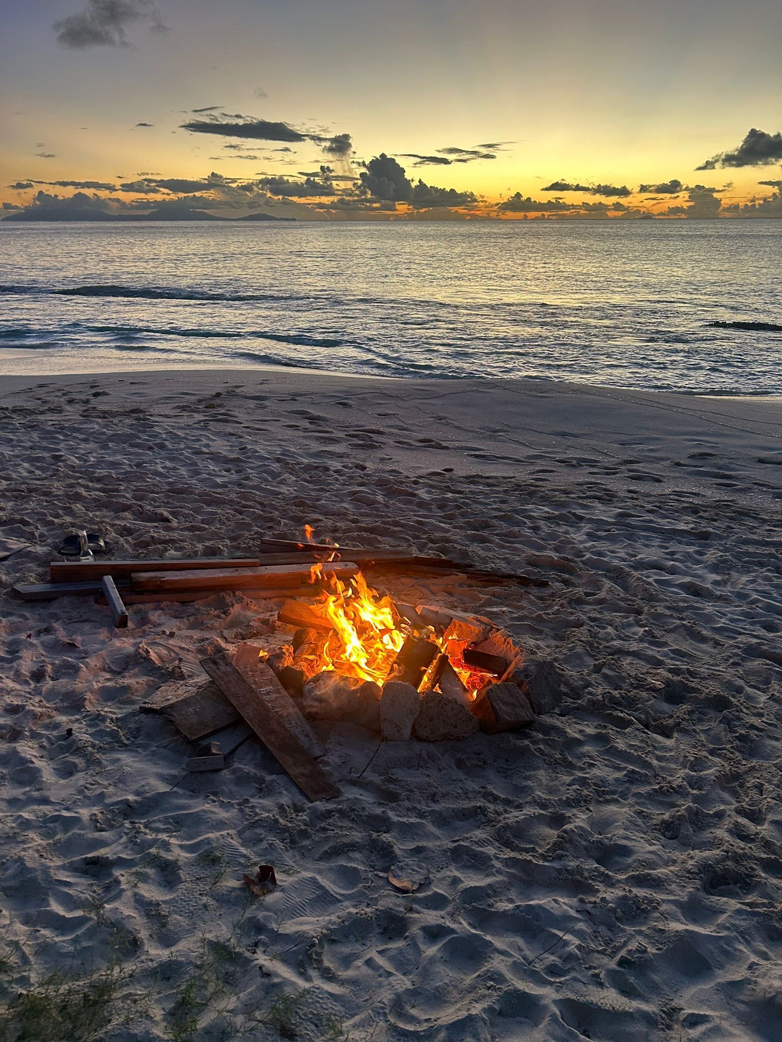 A beach scene with a fire pit in the sand, waves in the background, and a sunset.