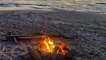A beach scene with a fire pit in the sand, waves in the background, and a sunset.