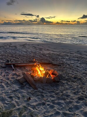 A beach scene with a fire pit in the sand, waves in the background, and a sunset. - Pool views meet the sea at Starfish House (Ffryes)
