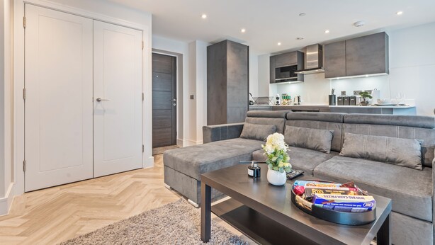 Een moderne woonkamer met een grijze bank, een salontafel met afstandsbedieningen en lekkernijen, en een keuken op de achtergrond.