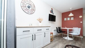 A modern kitchen with white cabinets, a clock, a flat-screen TV, and a dining area with white chairs and a table. - "Arzon" Apartment with Terrace, Shared Pool and Communal Garden (Arzon)