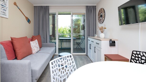 A modern living room with a sofa, a dining table, and a kitchen area with cabinets and a TV. - "Arzon" Apartment with Terrace, Shared Pool and Communal Garden (Arzon)