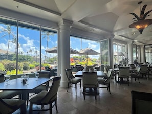 A restaurant with large windows, outdoor seating, and a view of palm trees and a garden. - Oceanview Studio at Kauai Beach Resort #2444 (Lihue)