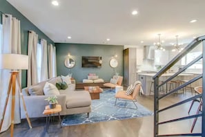 Ein modernes Wohnzimmer mit einer Couch, Couchtisch und Sesseln, ein Essbereich mit einer Küche im Hintergrund und einer Treppe, die zu einer weiteren Ebene führt.