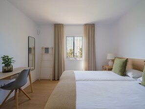 Ein Hotelzimmer mit Schreibtisch, zwei Betten, einem Fenster mit Vorhängen, einem Nachttisch mit Lampe und einer kleinen Pflanze.