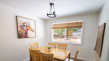 Un comedor con una mesa y sillas de madera, una lámpara colgante, un ventanal con persianas y un cuadro en la pared.