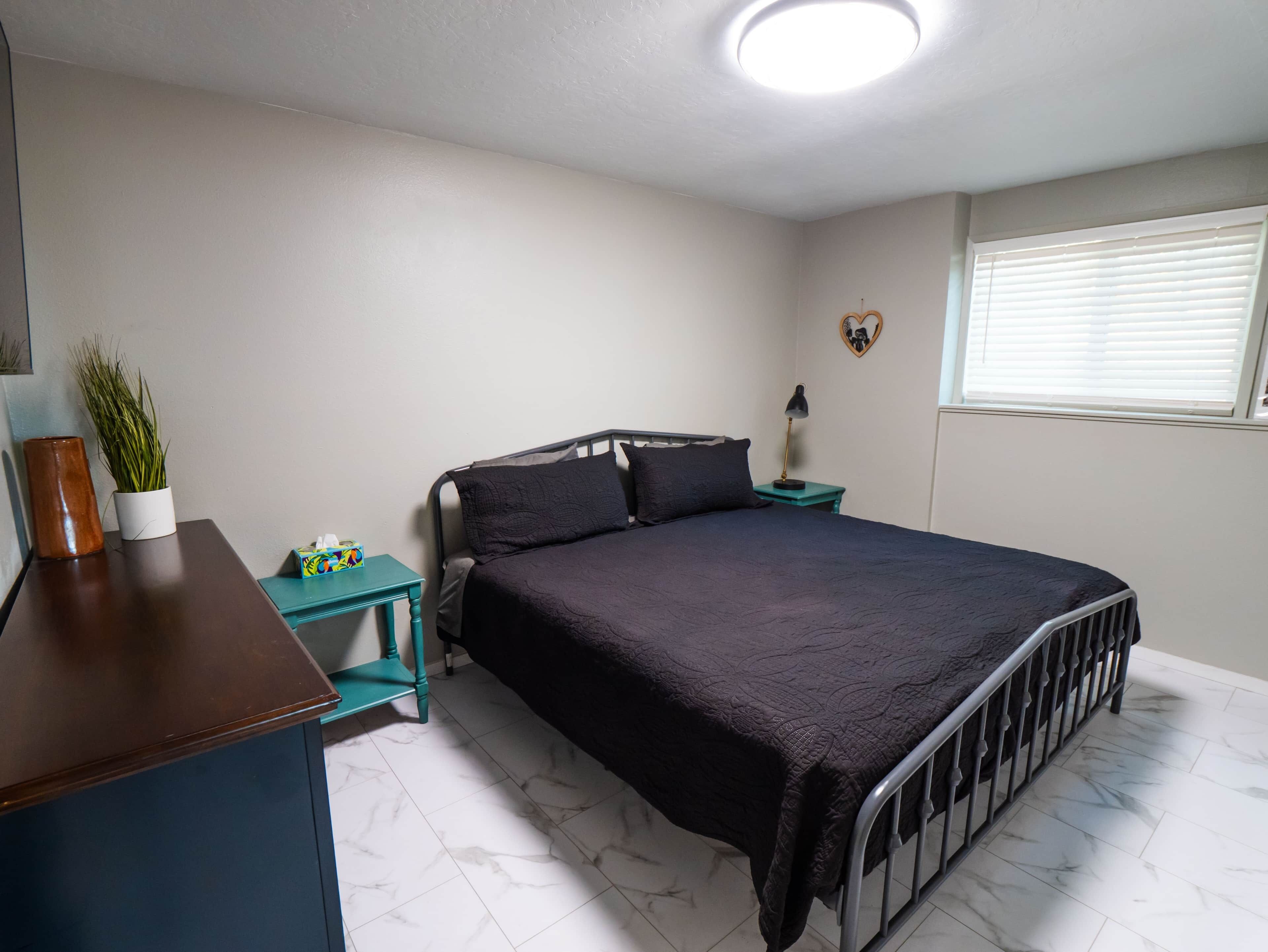 Ein Schlafzimmer mit Bett, Schreibtisch, Lampe, Topfpflanze und einem Fenster mit Jalousien.