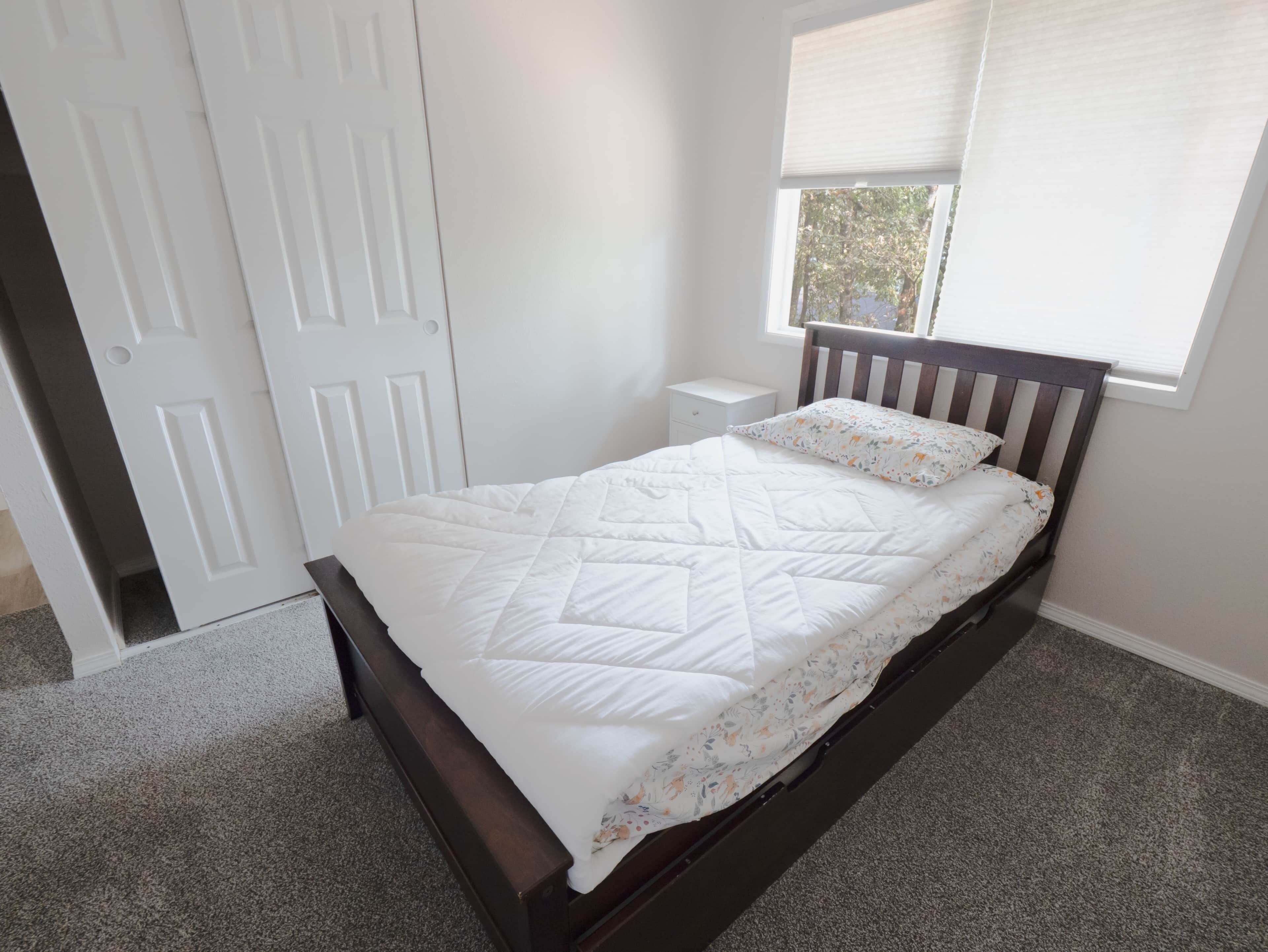 A bedroom with a single bed, a nightstand, a window with blinds, and a door.