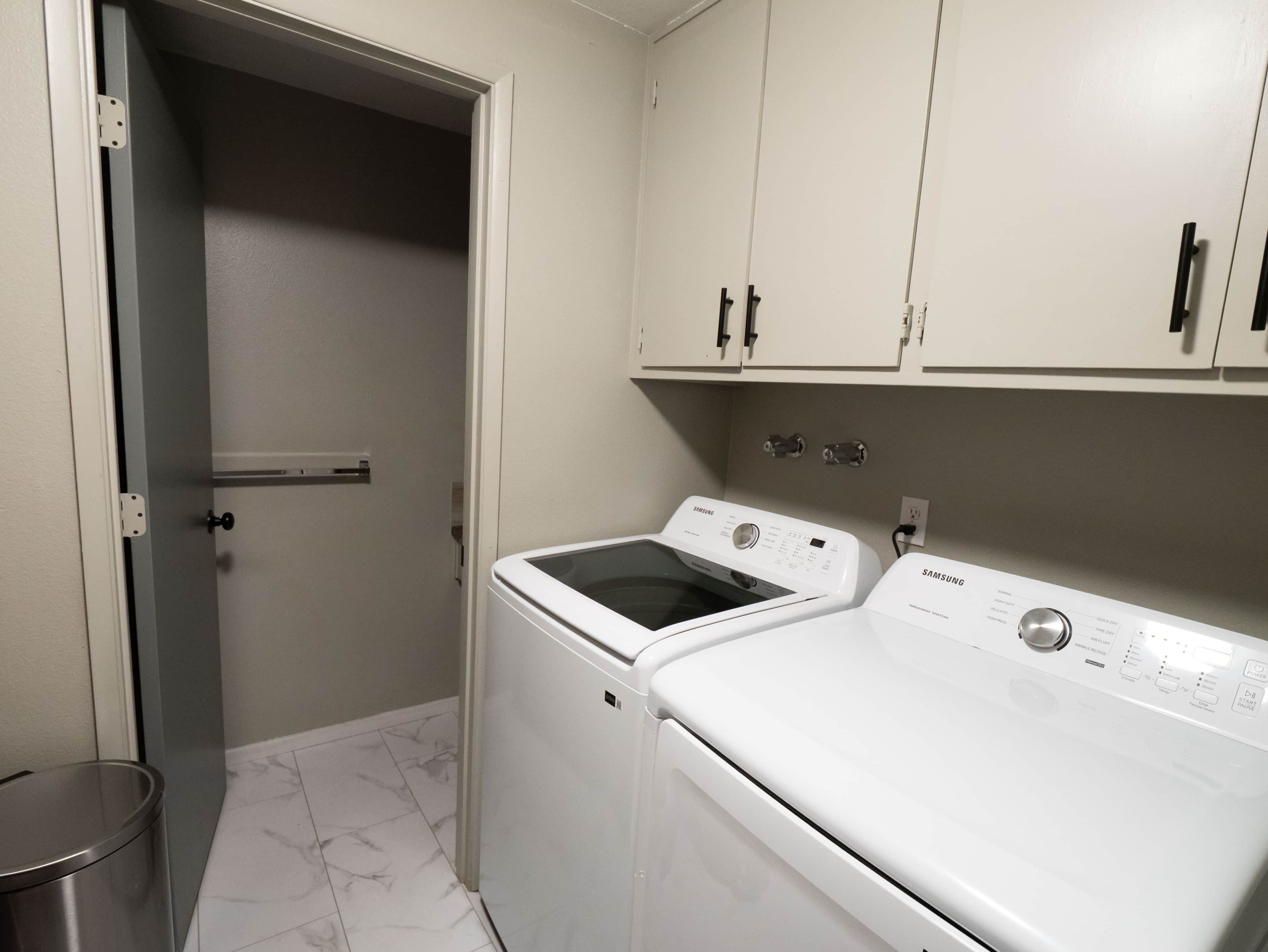 A laundry room with a washing machine and dryer, a trash can, and a door leading to another room.