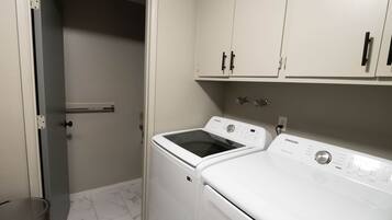 A laundry room with a washing machine and dryer, a trash can, and a door leading to another room.
