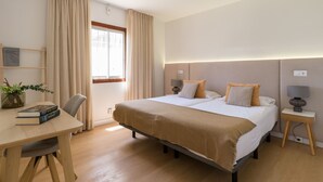 Habitación de hotel con cama, mesitas de noche, escritorio, silla, ventana con cortinas y una planta sobre el escritorio.