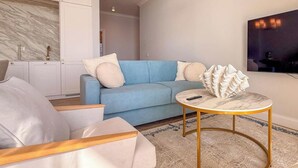Una sala moderna con un sofá azul, sillones blancos, una mesa de centro de mármol y una zona de cocina con armarios blancos.