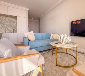 A modern living room with a blue sofa, white armchairs, a marble coffee table, and a kitchen area with white cabinetry. - Marlena - Corfu View Residence (Sarandë)