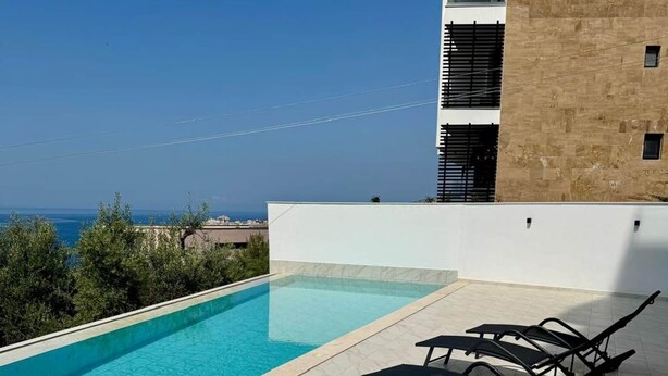 Una piscina en la azotea con vista despejada al mar, un edificio blanco y algunas sillas de descanso.