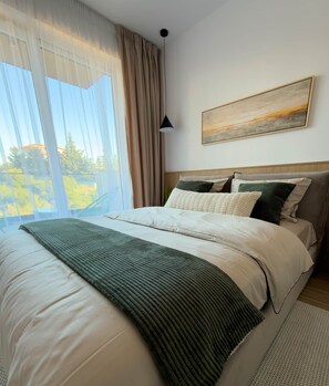 Een slaapkamer met een bed, gordijnen, een schilderij en uitzicht op groen.