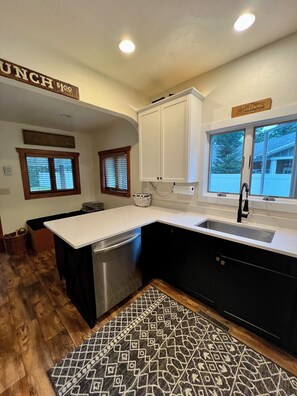 A kitchen with white cabinets, a stainless steel dishwasher, a patterned rug, and a window with blinds.