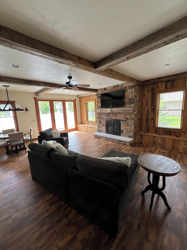 A spacious living room with a stone fireplace, wooden beams, a ceiling fan, and a large window.
