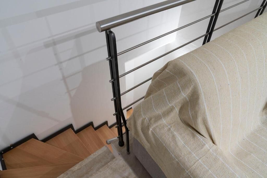 Una cama con estructura metálica y una colcha color beige, una escalera de madera y una pared blanca.