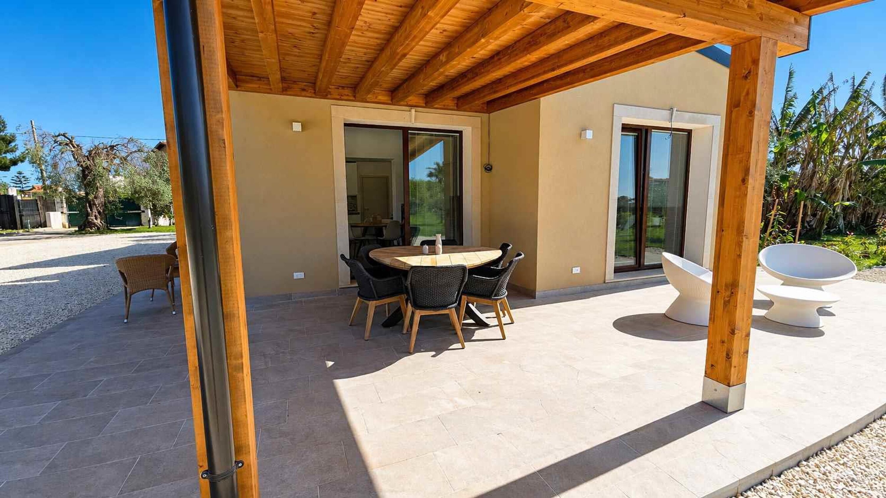 Eine überdachte Terrasse mit rundem Tisch und Stühlen, einer weißen Skulptur im Freien und Blick auf ein Wohngebiet mit Palmen.