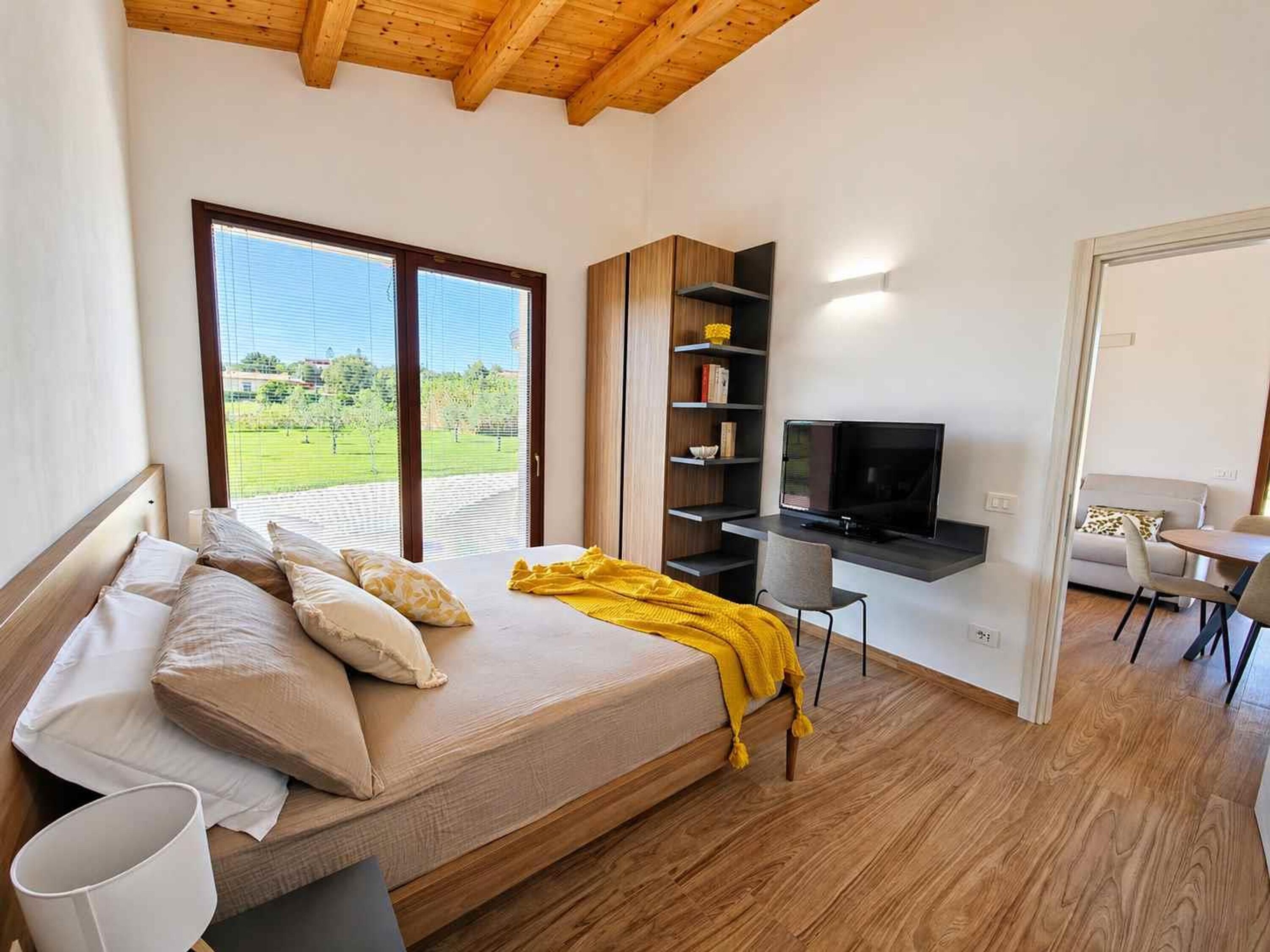 A bedroom with a bed, a desk, a television, and a view of the outdoors.