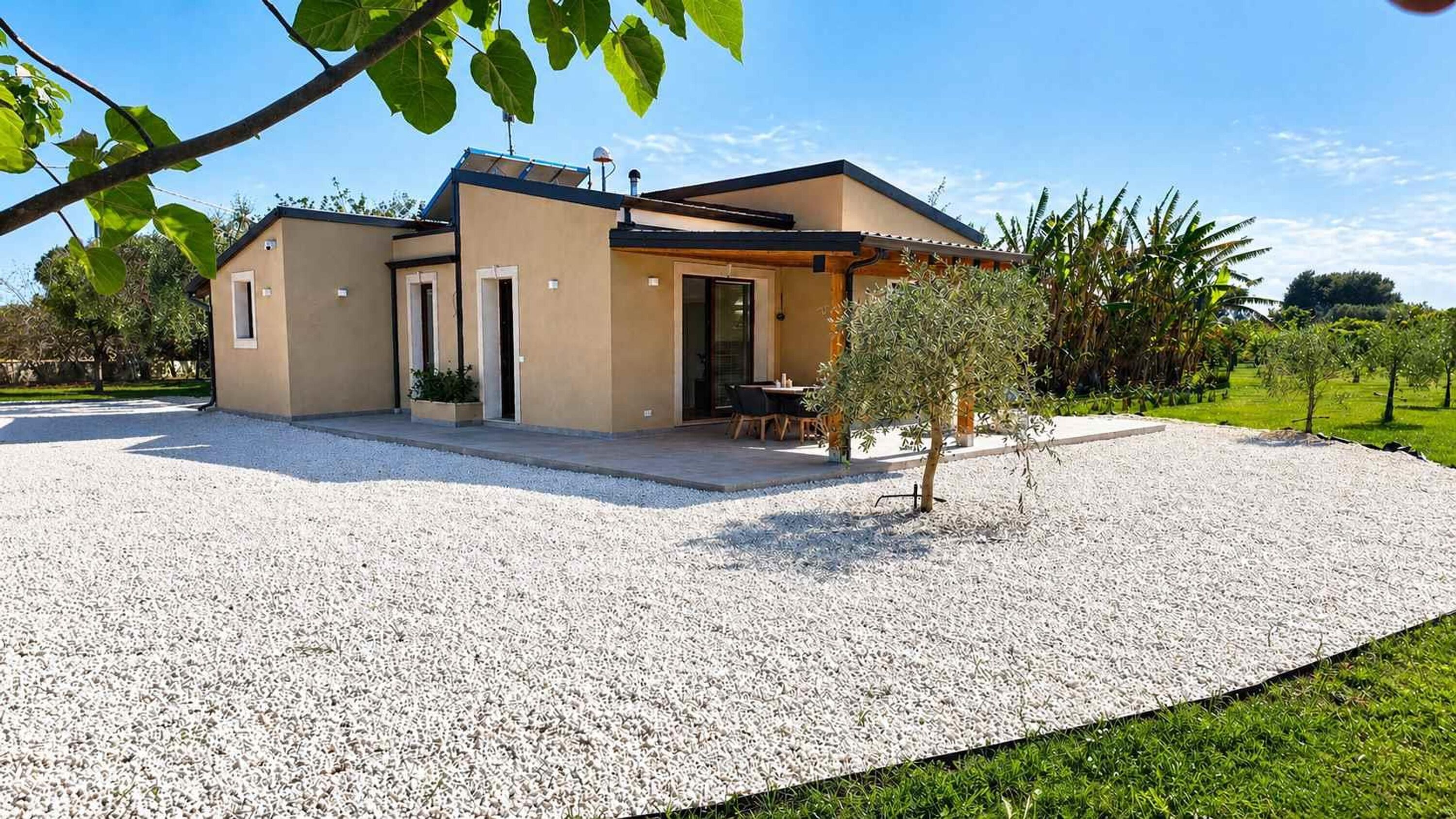 Una casa moderna con vialetto in ghiaia, patio con mobili da esterno e un piccolo albero.
