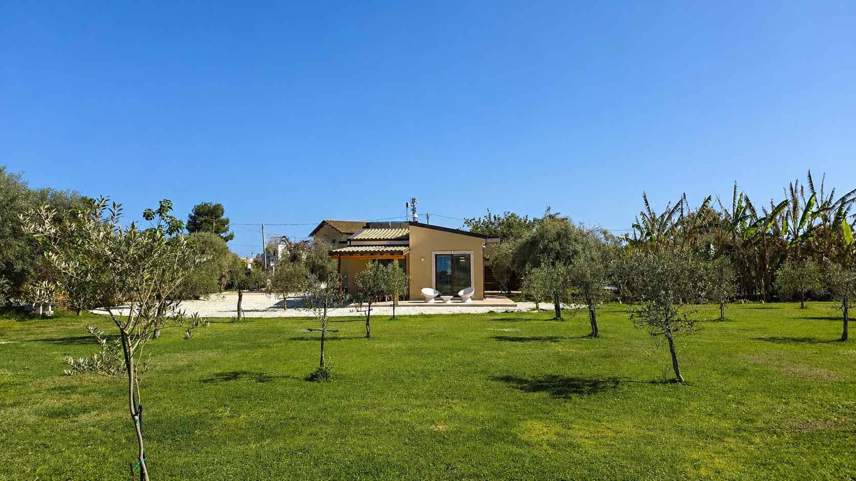 Una casa moderna con techo plano, un patio con dos sillas blancas y un césped bien cuidado con árboles jóvenes.