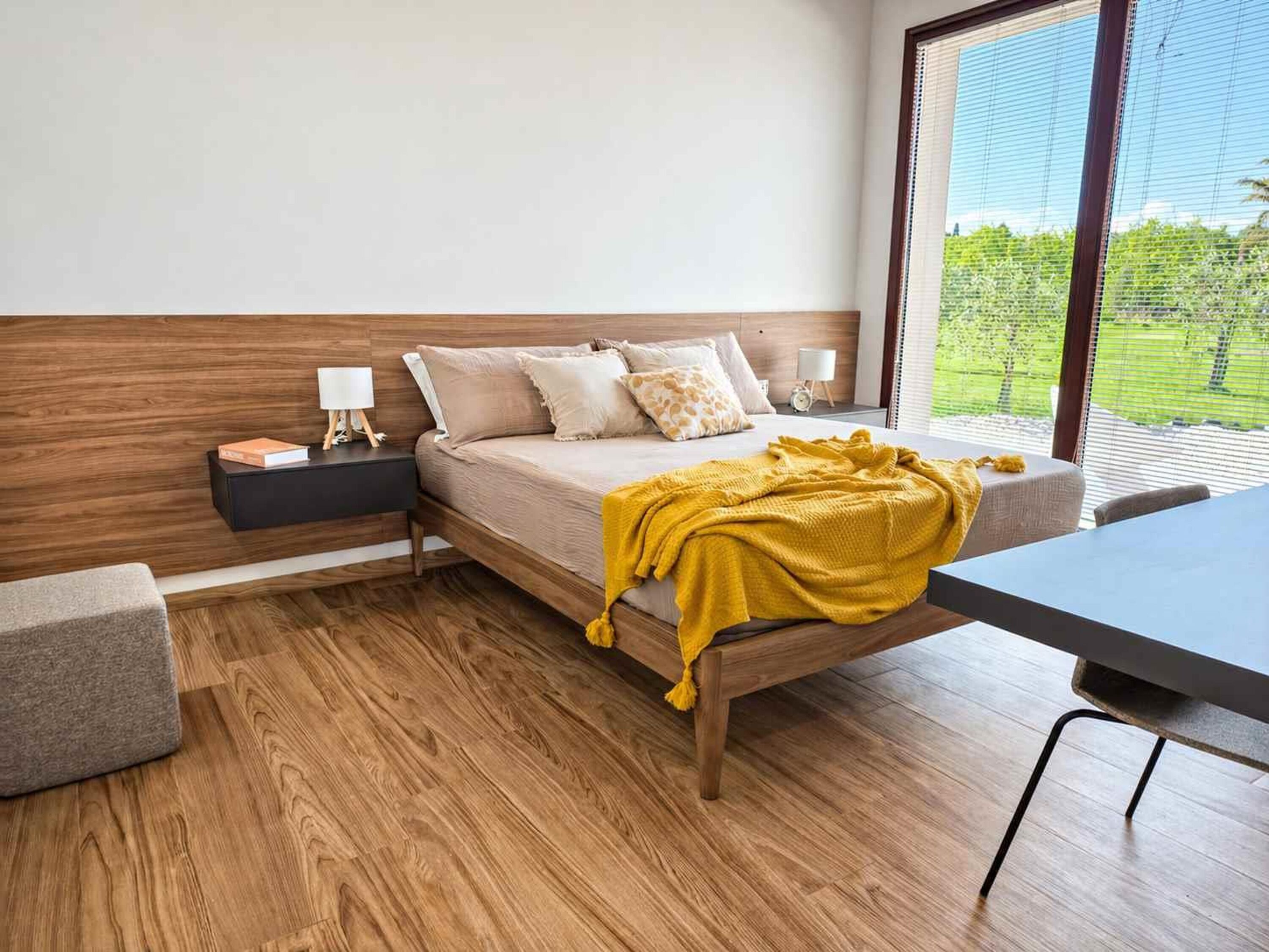 A modern bedroom with a wooden headboard, a bed with a yellow blanket, a bedside table with a lamp, and a view of greenery through the window.