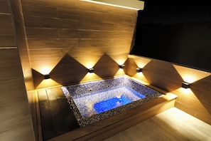 Un jacuzzi rectangular con base de mosaicos, rodeado de paredes de madera e iluminado por luces empotradas.