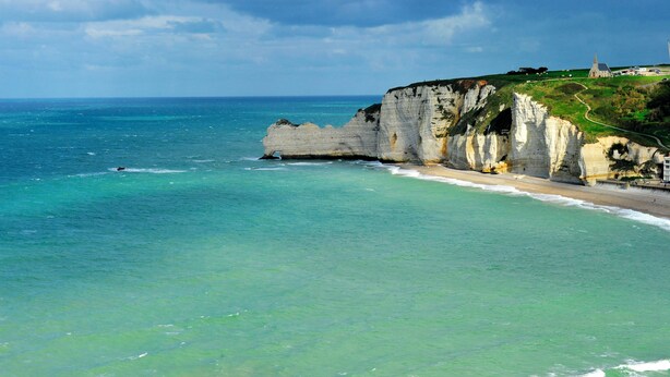 Paesaggio costiero con scogliere, una spiaggia e un sentiero che conduce a una costruzione sulla scogliera.