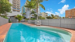 A swimming pool with a wooden deck, surrounded by a grey fence, with tall buildings in the background. - Canberra Heights Unit 5, 35 Canberra Terrace, King (Kings Beach)