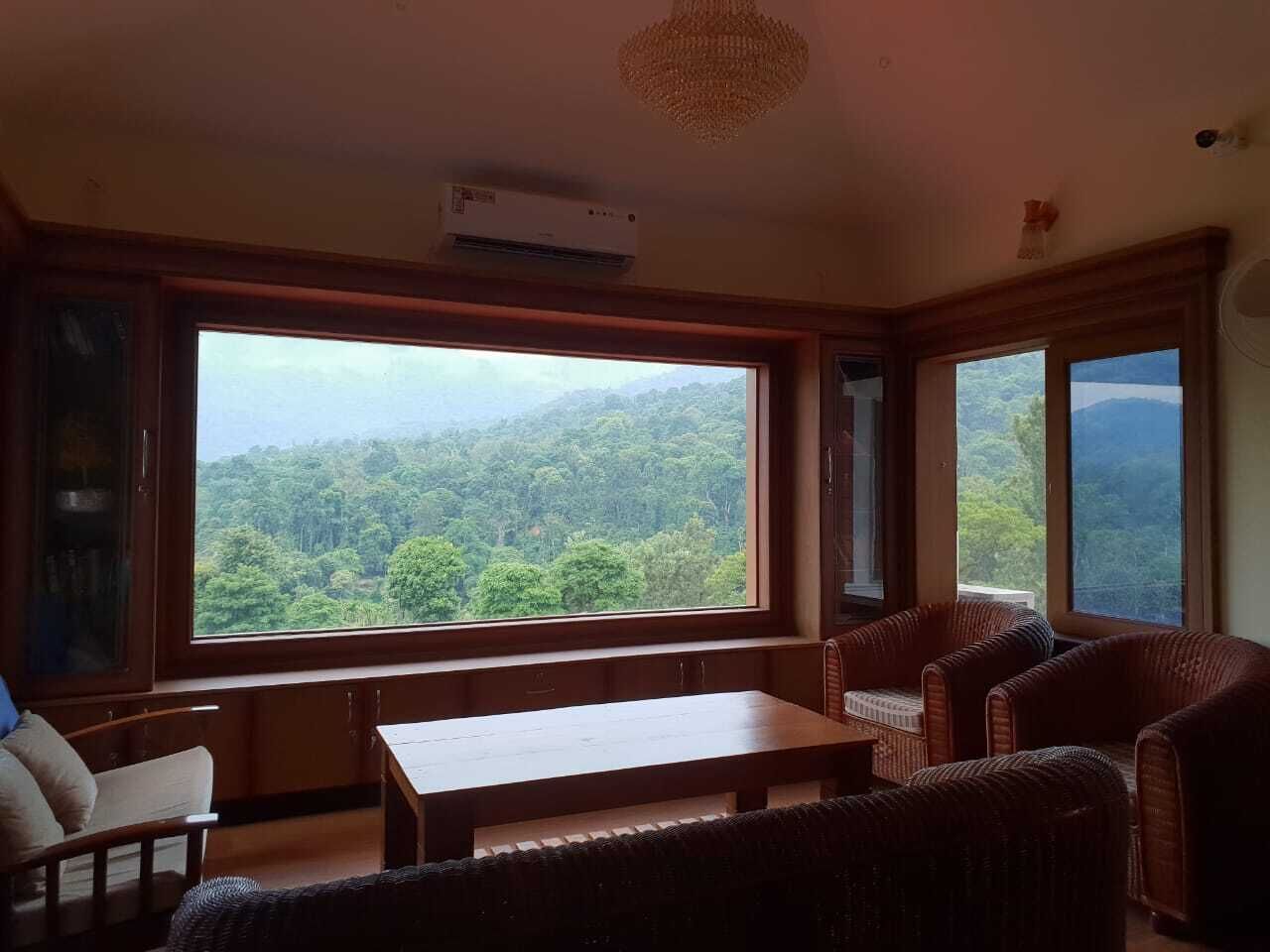 Ein Zimmer mit großem Fenster und Blick auf eine bewaldete Landschaft, einem Holztisch und Korbmöbeln.