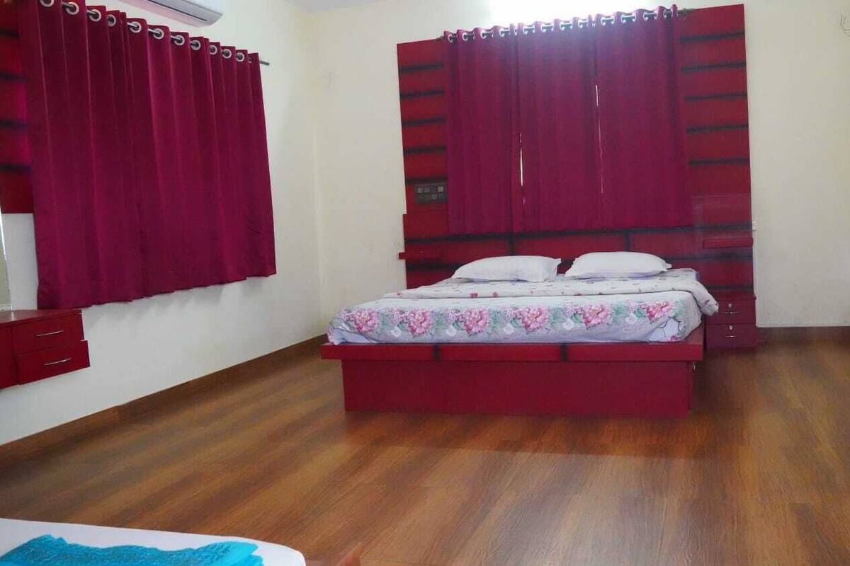 A bedroom with a wooden floor, a bed with a floral bedspread, red curtains, and a red headboard.