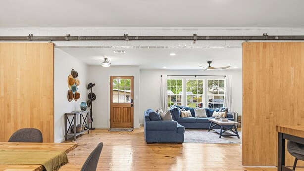A spacious living area with a wooden floor, a blue sofa, a dining table, and a large window with a view. - Girls Trip Getaway | Pool + Spa Near Main Street (Fredericksburg)