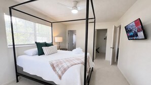 A four-poster bed with white bedding, a plaid throw blanket, and green pillows. There is a ceiling fan, a window with blinds, and a TV mounted on the wall. - Heated pool home with a lake view! (St. Augustine)