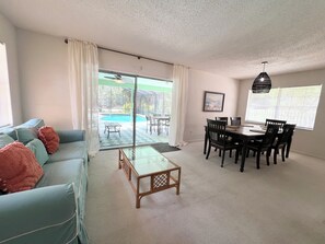 Una sala de estar con sofá, mesa de centro, mesa de comedor, sillas y vista a una piscina.