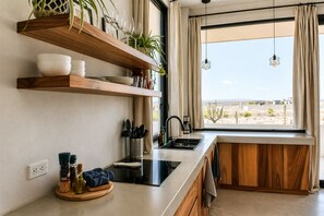A modern kitchen with wooden shelves, a black countertop, and a window with a view of a desert landscape. - Ocean view terrace and saltwater pool group getaway (Todos Santos)
