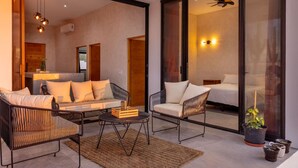 Um interior moderno com sofá, poltrona e uma pequena mesa. Há uma cama com cabeceira e um ventilador de teto.