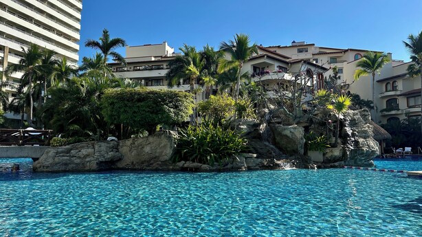 Una piscina azul cristalina con una isla rocosa en el centro, rodeada de palmeras y edificios.