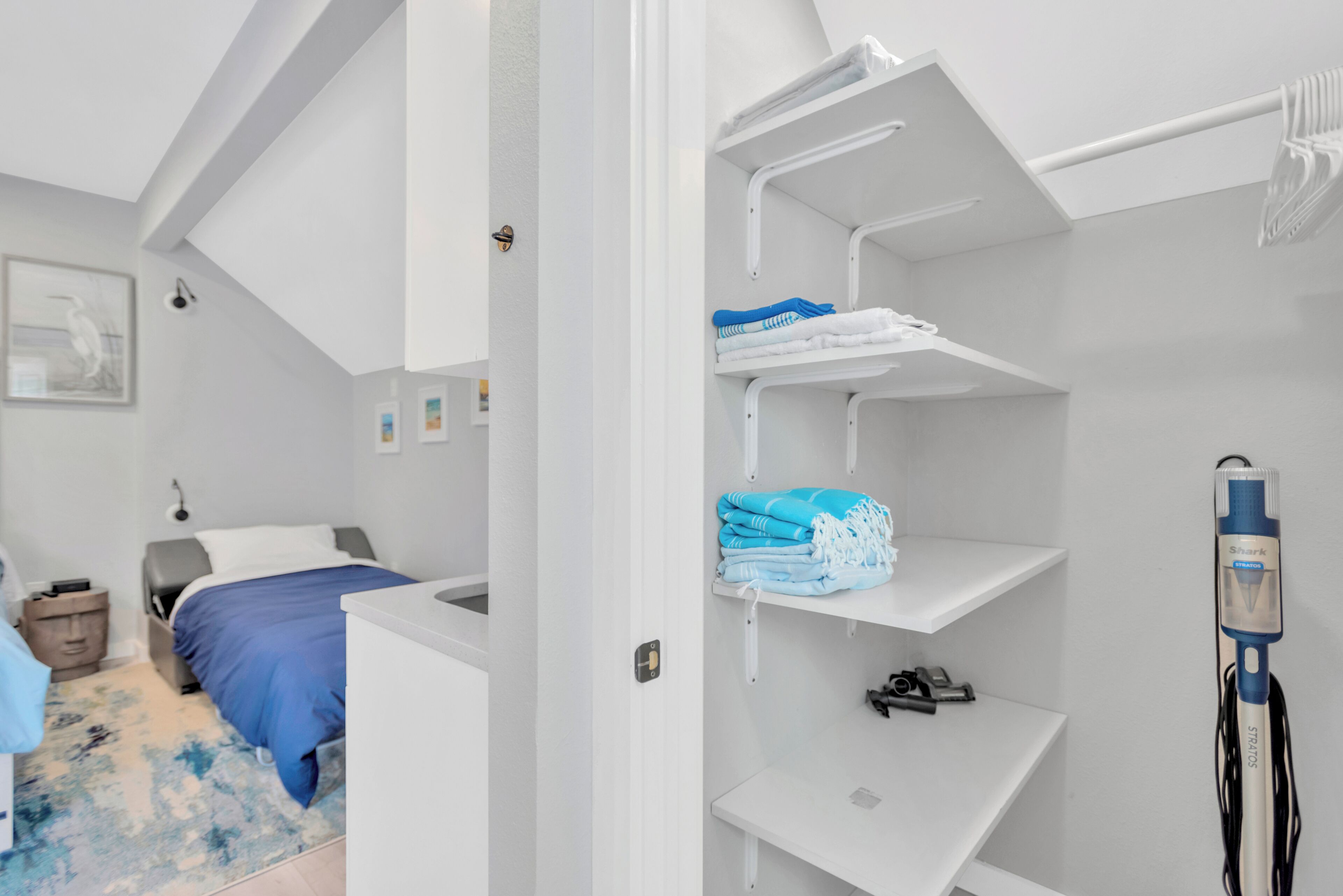 Un dormitorio compacto con cama, un escritorio pequeño, un armario blanco con estantes y una mesita de noche.