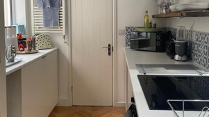 A compact kitchen with a white door, a window with a blue curtain, a microwave, a kettle, and a coffee machine. - Gorgeous One Bedroom Flat in Woolwich Arsenal (London)