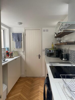 A compact kitchen with a white door, a window with a blue curtain, a microwave, a kettle, and a coffee machine. - Gorgeous One Bedroom Flat in Woolwich Arsenal (London)