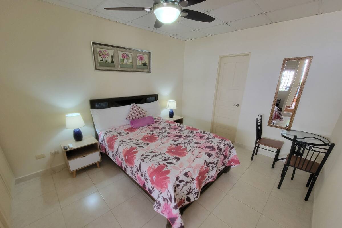 A bedroom with a bed, bedside lamps, a ceiling fan, a desk with a chair, and a mirror.