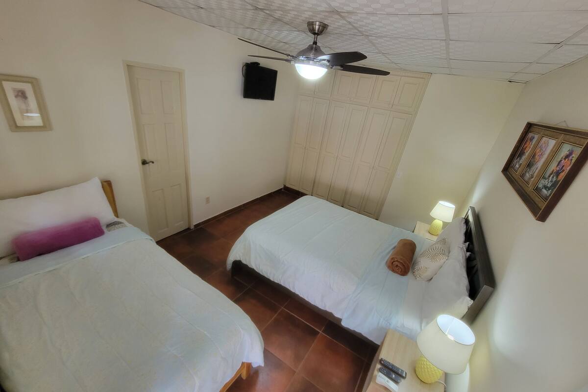 A hotel room with two beds, a ceiling fan, a TV, and a small table with a lamp.