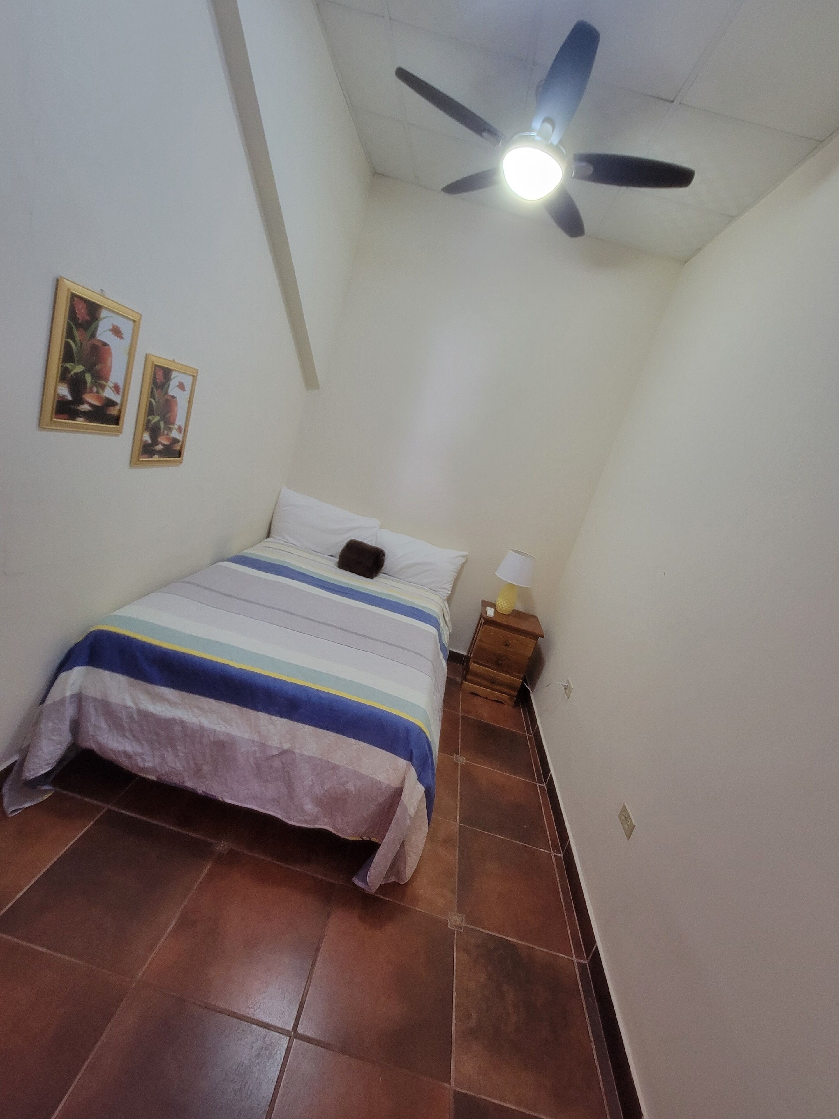 Ein Schlafzimmer mit einem Bett, einem Deckenventilator, einem Nachttisch und zwei gerahmten Bildern an der Wand.